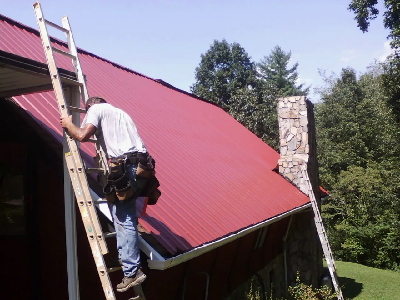 Roofer on a ladder beside a red metal roof and stone chimney — Soffit & Fascia Repair by Roofing Brothers in Crewe