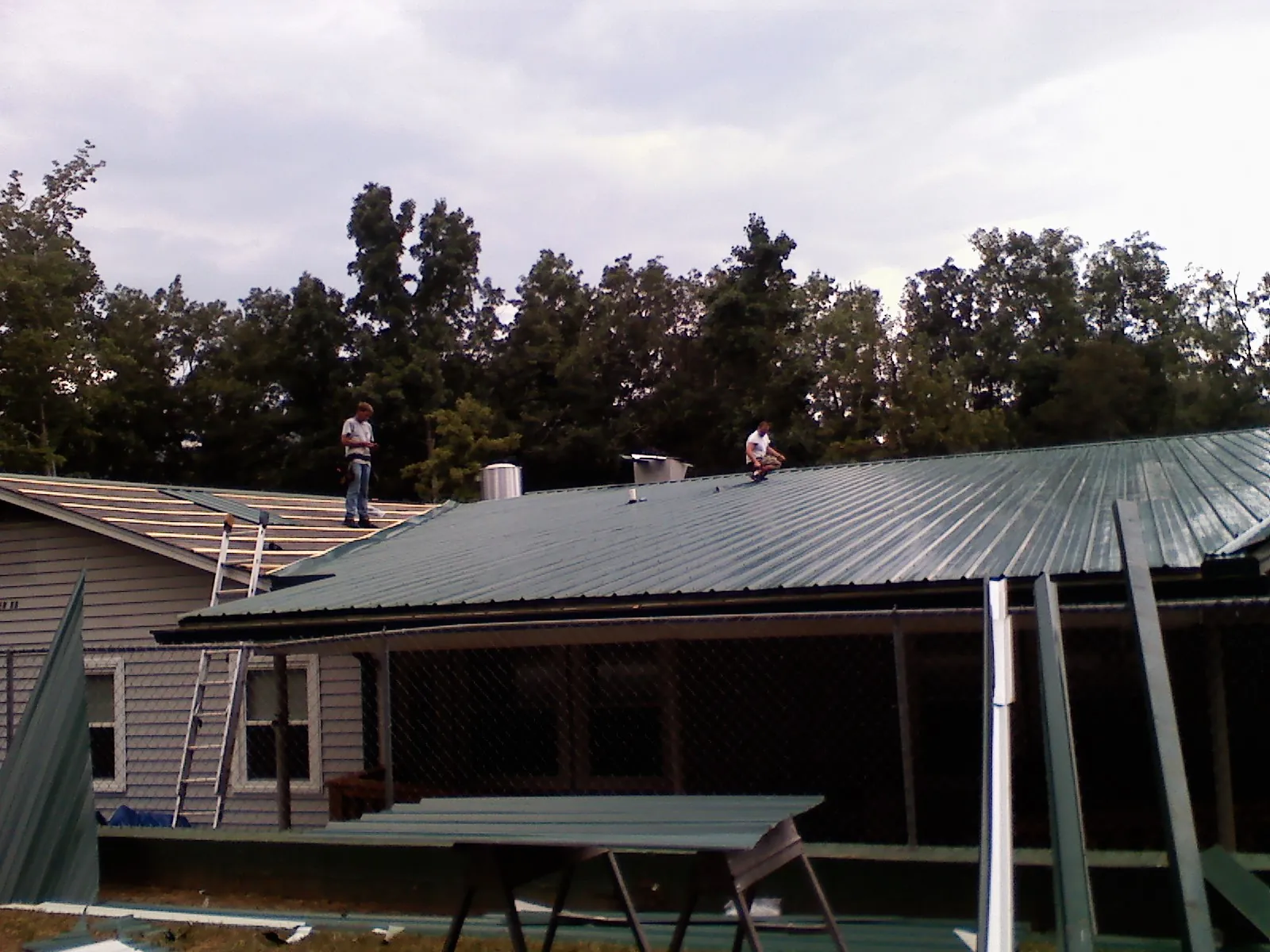 Two roofers installing green metal panels above a covered porch in Crewe